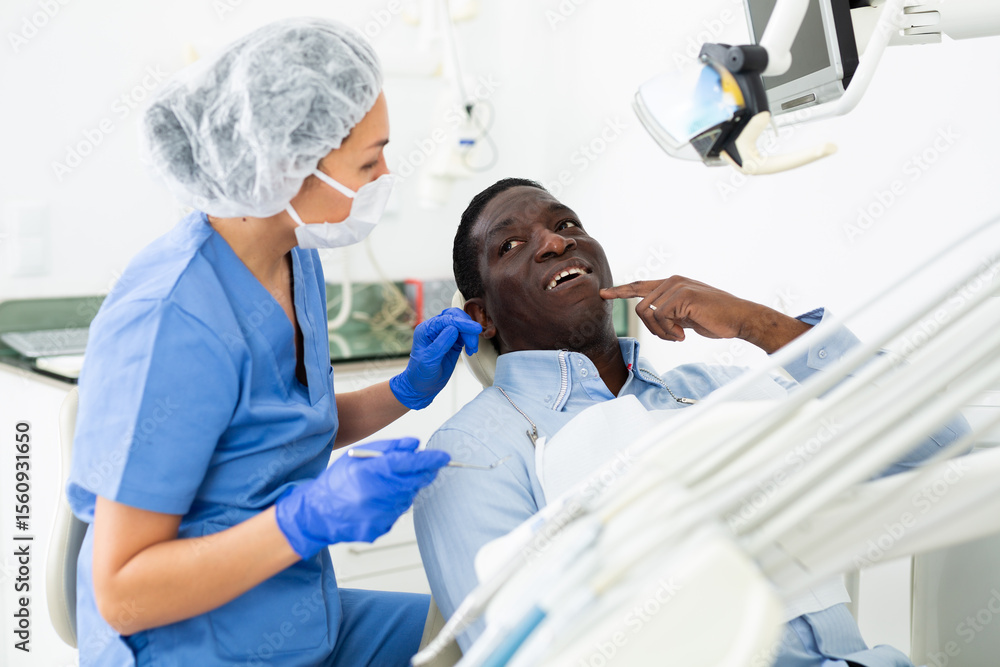 Obraz premium African american patient talking to female dentist and complaining about toothache at dental clinic office..
