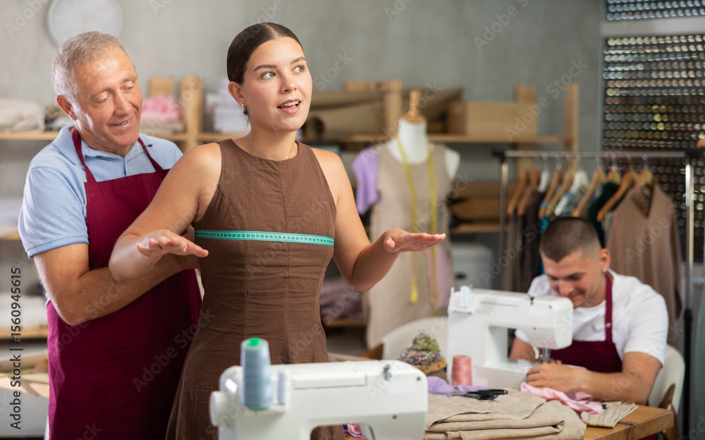 Obraz premium Elderly tailor takes measurements from a woman to sew clothes according to an individual sketch. Young employee of the atelier works on a sewing machine
