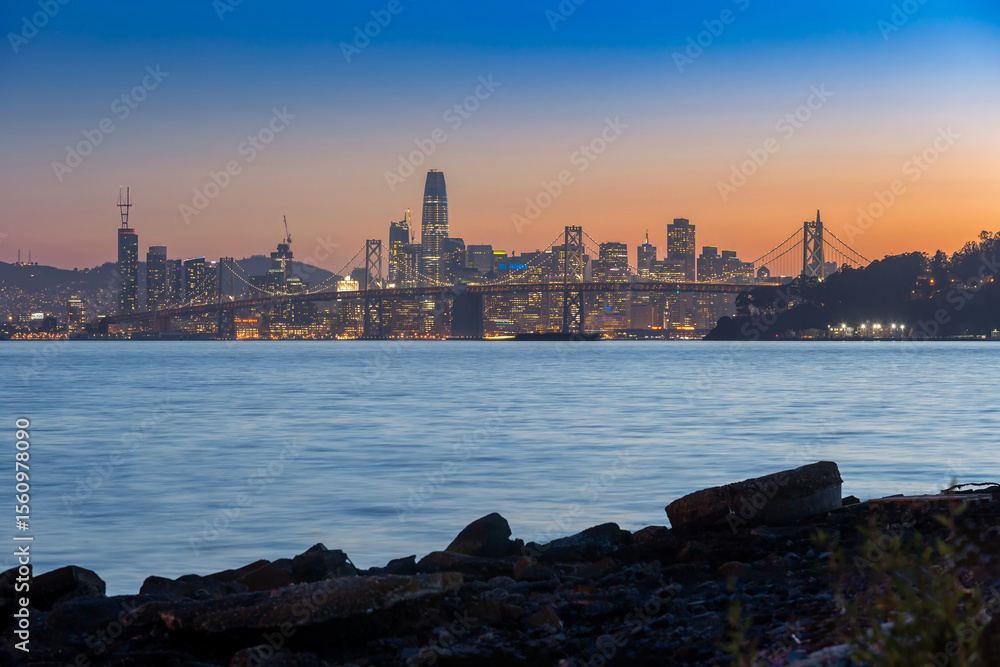 Fototapeta premium San Francisco, USA: The city skyline and Bay Bridge are illuminated at dusk. The lights of the buildings and bridge reflect on the water.
