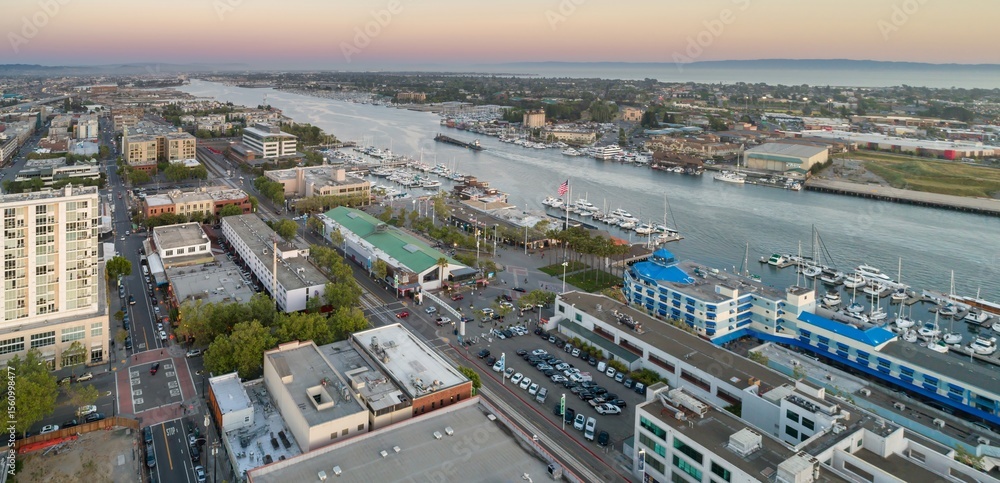 Fototapeta premium Aerial view of ack London Square, Oakland, California, USA, showing the Oakland-Alameda Estuary, buildings, parking lots, and streets at dusk. The image captures the urban landscape.