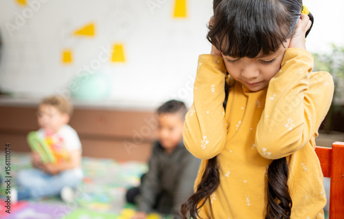 Preschool girl covers her ears and closes her eyes while other kindergarten children play loudly behind her. Concept of noise sensitivity, sensory overload, and emotional response in early childhood