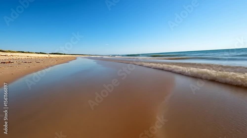 Coastal scene depicting a sandy beach with gentle waves under a bright blue sky. The water reflects the sky. The sand is wet and has a unique texture. A scenic view of the coastline