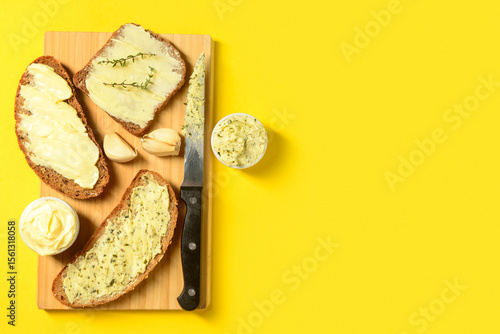 Tablou pe pânză Wooden board of bread slices with fresh butter and garlic on yellow background