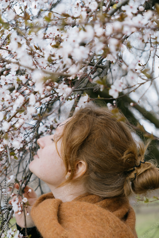 Naklejka premium woman with red hair stands in front of a tree with pink flowers. She's wearing a brown coat, scarf.