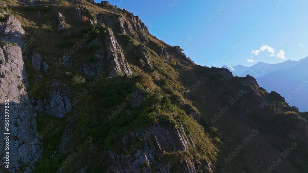 Aerial shot of a mountain slope with rocky cliffs in Kyrgyzstan, camera moving right showing distant snow-covered peaks on the horizon at sunny evening with clear blue sky. Aerial shot of a mountain slope with rocky cliffs in Kyrgyzstan, camera moving right showing distant snow-covered peaks on the horizon at sunny evening with clear blue sky.