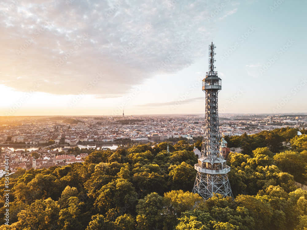Obraz premium Golden light bathes Petrin Lookout Tower as the sun rises over Prague. Surrounded by lush greenery and the city skyline, this moment captures the beauty of early morning tranquility.
