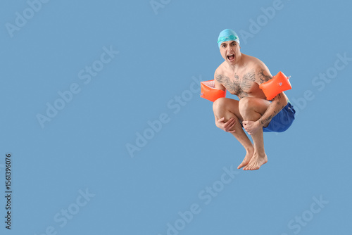 Handsome male swimmer with inflatable armbands doing cannonball jump on blue background