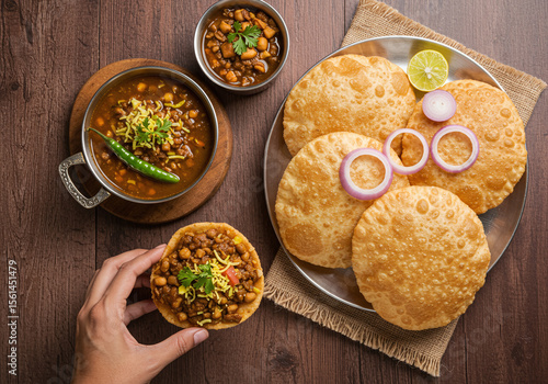 Spicy Indian chole curry served with crispy puri bread and garnished with fresh coriander, onion rings, and lime on wooden table, showcasing traditional vegetarian cuisine
