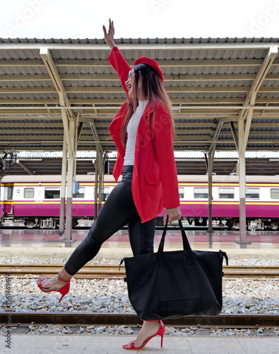 Joyful Asian woman wears a red coat, a matching red beret, black pants, and red high-heeled shoes at a train station platform