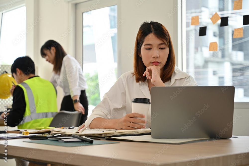 An Asian Female engineer thoughtfully working on a laptop while holding a coffee cup. Colleagues collaborate in the background in a bright engineering office An Asian Female engineer thoughtfully working on a laptop while holding a coffee cup. Colleagues collaborate in the background in a bright engineering office