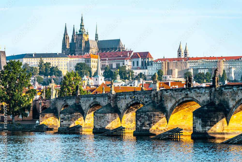Obraz premium Early morning light casts a golden hue on Charles Bridge as it arches over the Vltava River. Prague Castle looms majestically in the background, surrounded by historic architecture.