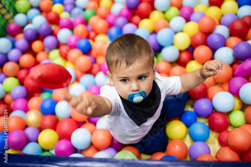 Baby boy playing with colorful balls in ball pit: throwing red ball and having fun