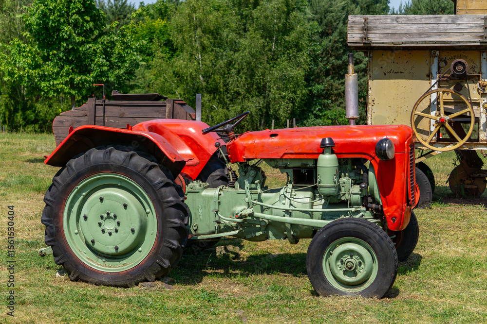 Obraz premium Classic red and green vintage tractor parked on grass, with visible engine details and large rear wheels. Surrounded by rural scenery and old agricultural machinery