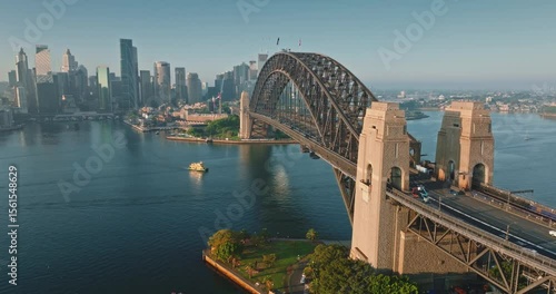 Australia, Sydney: Aerial view Sydney Harbour Bridge with cars riding in morning light, a ferry sails below. Travel destination, modern cityscape architecture in background. Drone footage panorama