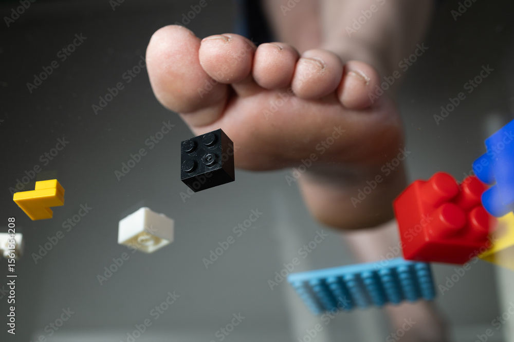 Naklejka premium A foot is shown stepping on colorful construction toy bricks scattered on the floor, highlighting the chaos of playtime. The setting suggests a fun but messy environment created by children.