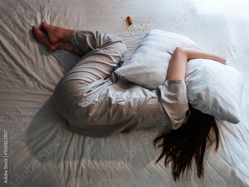 woman lying on bed, cover her head with pillow suffer from insomnia, sleeping pills next to her on bed. top above view of woman curl up in stress in bedroom with sleep disorder