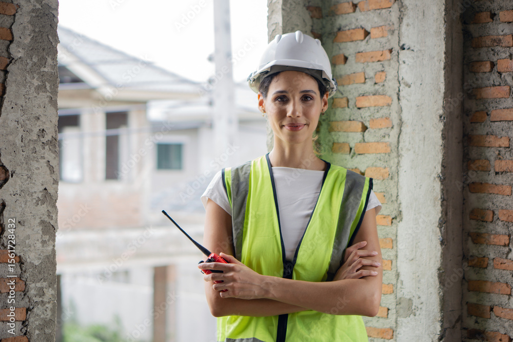 Fototapeta premium Female construction worker in hardhat and safety vest stand confidently with arms crossed in a partially built structure while holding a walkie-talkie during daytime.