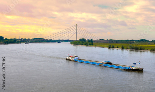 Editorial image of a cargo ship sailing on the Rhine River in Wesel, Germany, with the Niederrhein Bridge visible in the background. Shot on 30 May 2024.