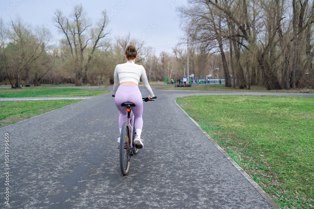 Fototapeta premium Young woman riding a bicycle in the park on the background of trees. Trendy active girl spends time on the nature. Side view.