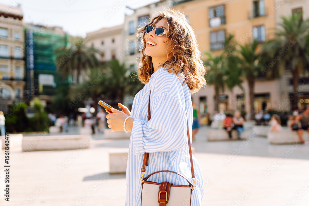Obraz premium A joyful young woman in sunglasses and holding a phone in her hands walks through sunny streets. A happy tourist with a phone enjoys new architecture on a sunny day. Tourism and enjoyment concept.