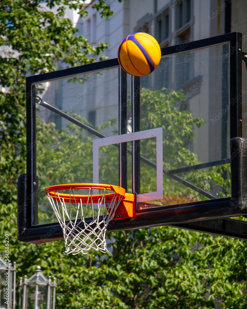 Naklejka premium Yellow and purple basketball flying toward the hoop on a clear sunny day. Urban outdoor sports scene with focus on street basketball and active lifestyle.