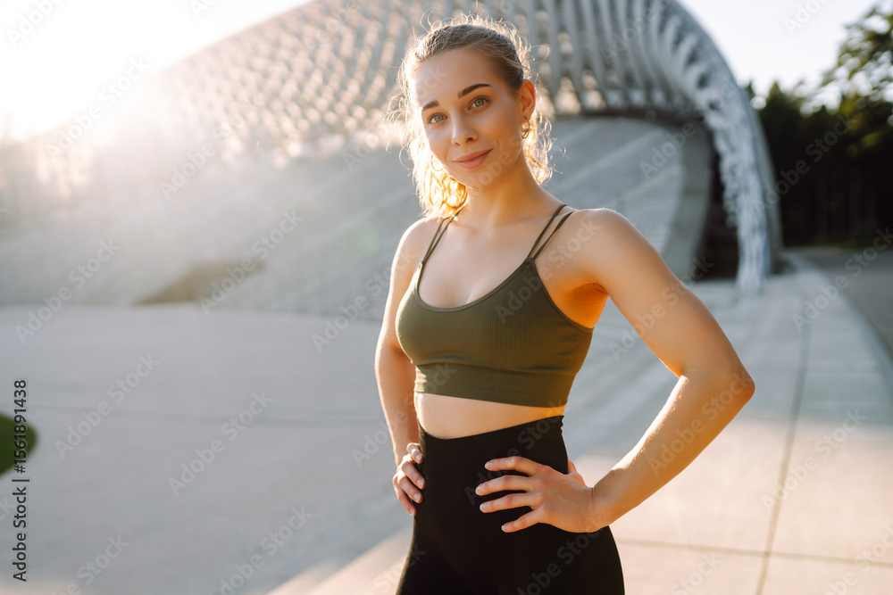 Fototapeta premium Portrait of a happy woman in sportswear standing in the rays of the sun on a sports ground. The athlete is warming up and enjoying the sunny weather outdoors. Sports concept. Active lifestyle.
