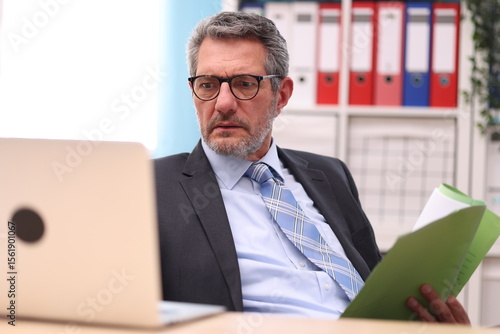 Businessman in black suit looks very concentrated into his laptop with a file in his hand