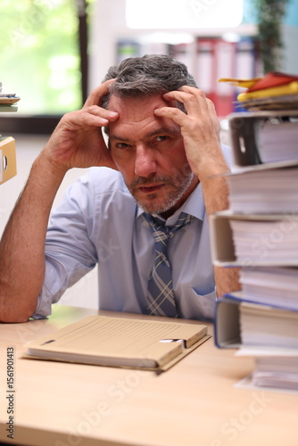 Office worker in shirt and tie sitting in the office in front of stacks of files and looks very annoyed and exhausted