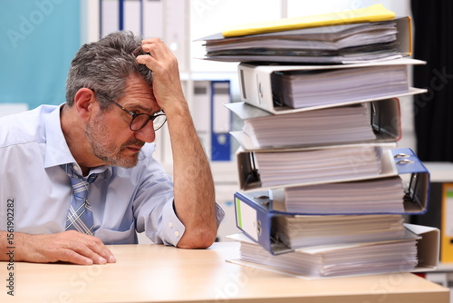 Office worker with glasses sits at the desk, supports his head with one arm and looks depressed to the side