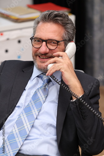 Office worker with glasses and suit sits at his desk, talks on the phone and is very satisfied with the conversation