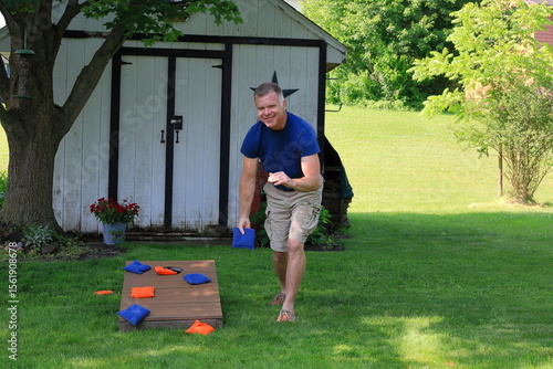 Middle aged man in a blue shirt is throwing corn hole bags during a backyard game