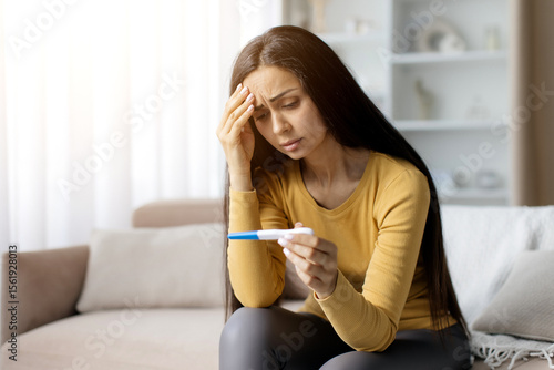 Fotografía Worried young woman sitting on sofa, holding positive pregnancy test with hand o