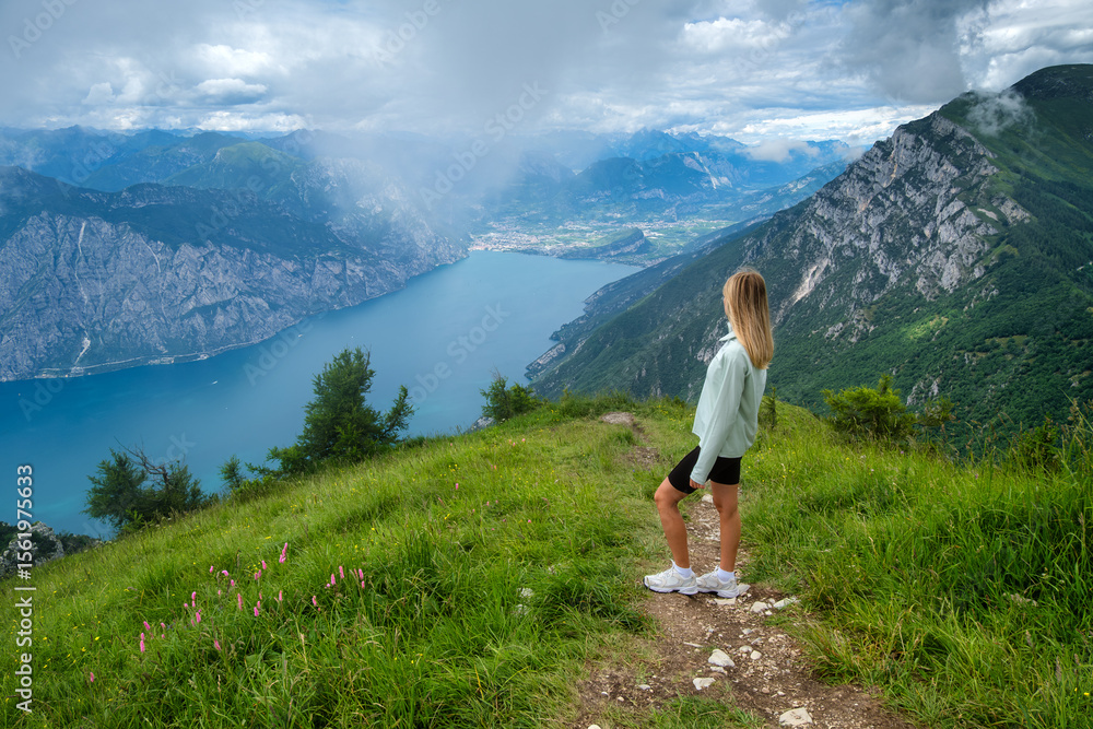 Naklejka premium Woman tourist standing on a mountain path, admiring the stunning view of Lake Garda, surrounded by lush vegetation and a cloudy sky in summer. Travel destination. Girl and Alpine lake in Italy