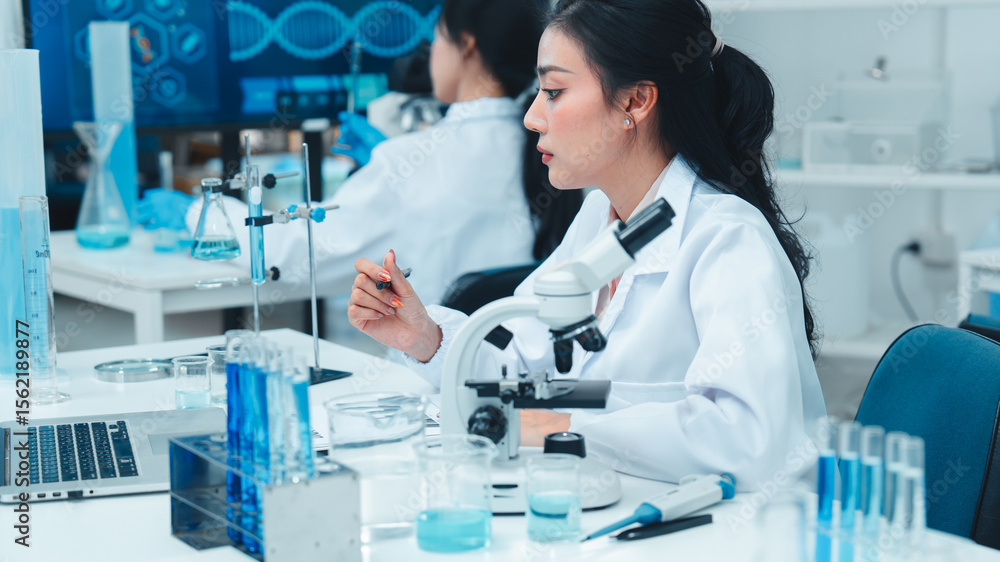 Asian female scientist in lab coat working on a laptop and taking notes in a modern laboratory. Scientific research environment with test tubes, beakers, and microscope in the background. Asian female scientist in lab coat working on a laptop and taking notes in a modern laboratory. Scientific research environment with test tubes, beakers, and microscope in the background.