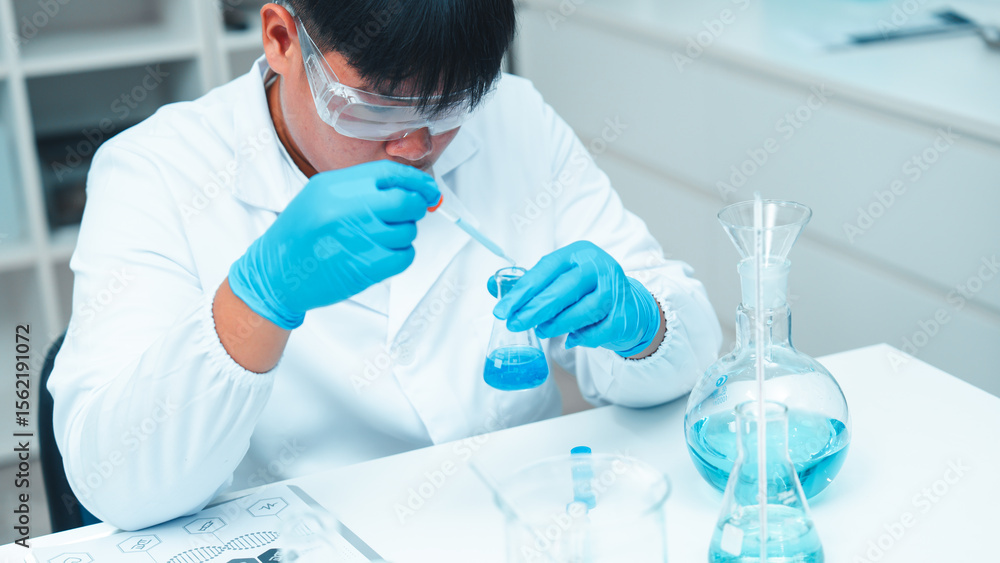 Fototapeta premium Asian male scientist wearing lab coat and safety goggles using pipette to transfer blue liquid in modern laboratory. Beakers and flasks with chemical solutions are on the table.