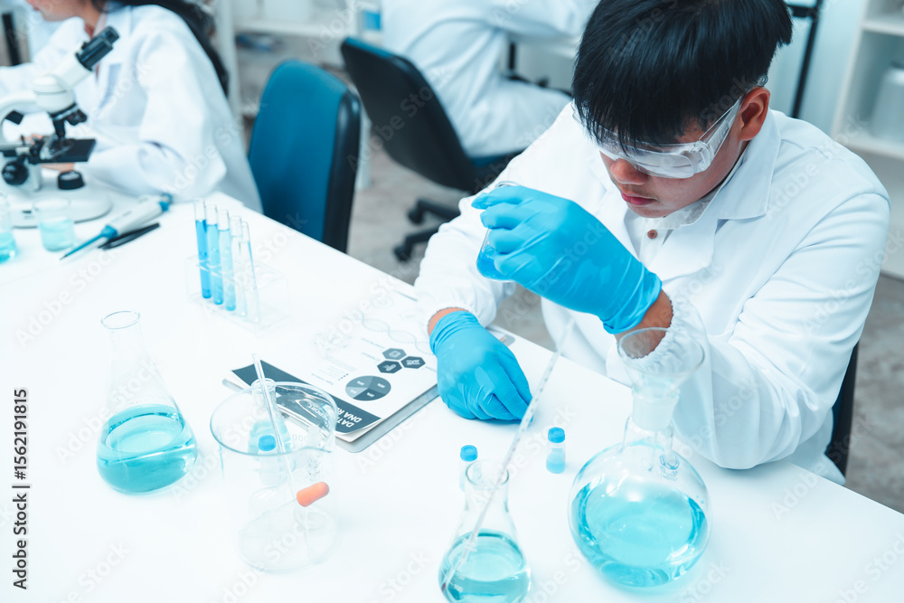 Fototapeta premium Asian male scientist wearing lab coat and safety goggles using pipette to transfer blue liquid in modern laboratory. Beakers and flasks with chemical solutions are on the table.