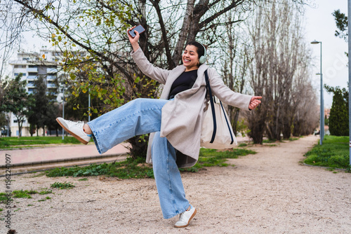 Young stylish influencer dancing and listening to music with headphones and smartphone in a park
