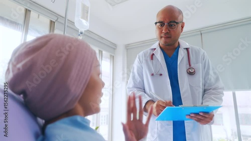 Asian woman patient describes her symptoms to an oncologist while sitting on a bed and looking at a man doctor in a lab coat in a hospital ward, health and medical care concept