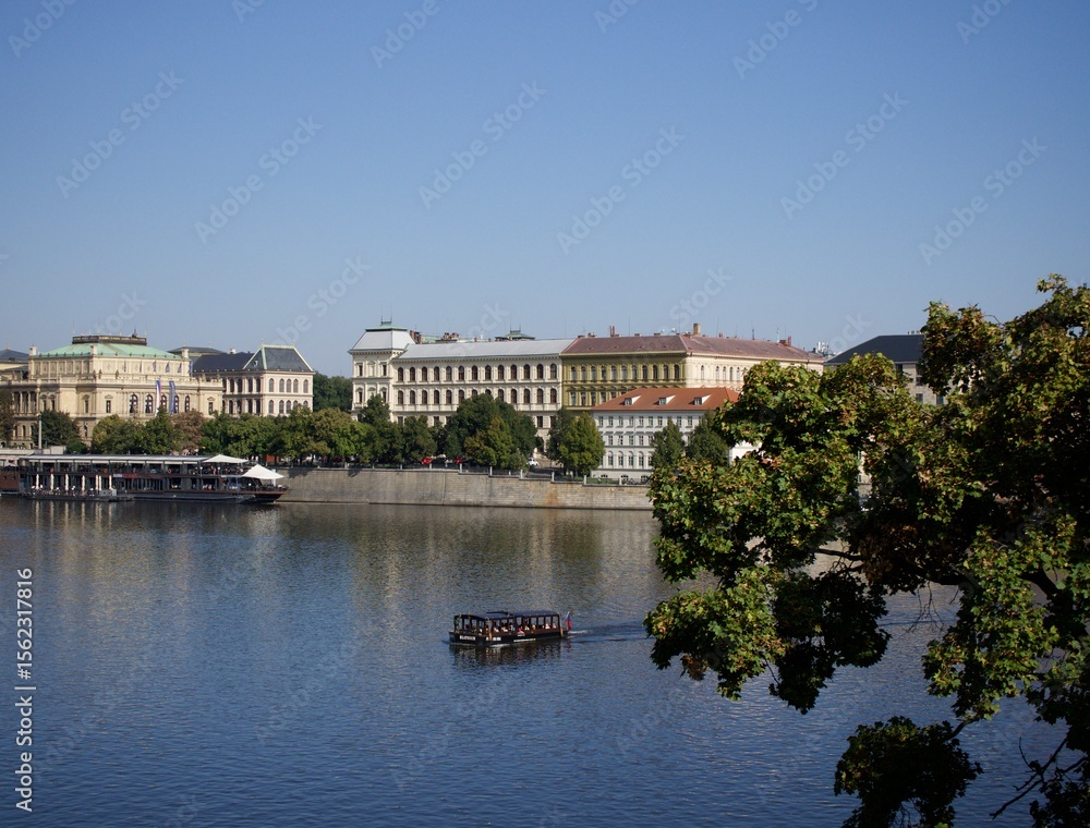 Naklejka premium Serene waters of a European city reflecting historic architecture under a clear sky, with a boat cruising by.