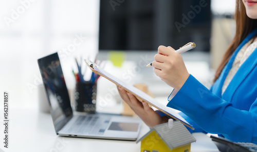 Tableau sur toile Property Professional at Work: A real estate professional meticulously reviews documents at her desk, laptop and a miniature property model, illustrating diligence and dedication in real estate