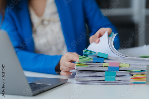 Workplace Documentation: A close-up shot of an office worker in a tailored blue blazer, meticulously reviewing a stack of documents.