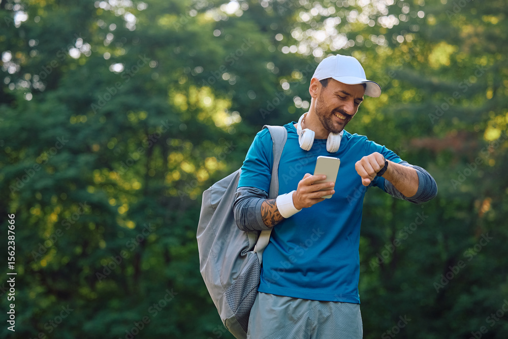Obraz premium Happy sportsman using smart watch and cell phone while exercising in nature.