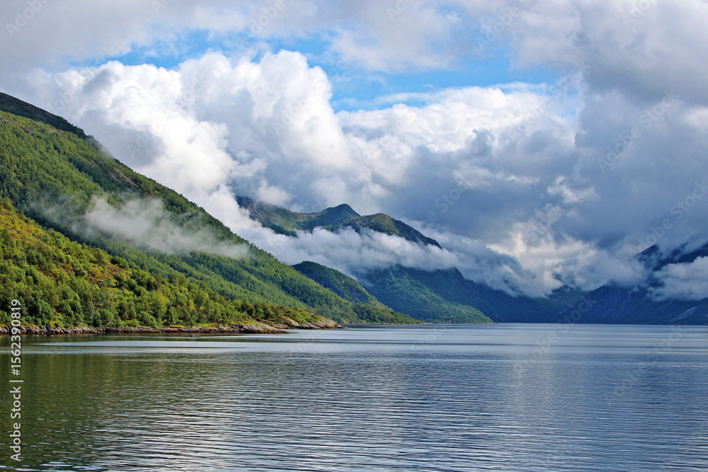 Calm waters reflect majestic mountains and vibrant greenery under a vivid sky with fluffy clouds in the distance Calm waters reflect majestic mountains and vibrant greenery under a vivid sky with fluffy clouds in the distance