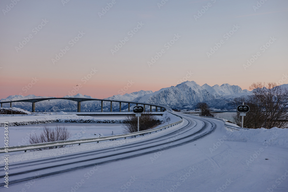 Snowy road curving toward bridge and mountain range at sunset Snowy road curving toward bridge and mountain range at sunset