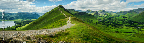 Panoramic landscape of Catbells fell with a visible hiking trail leading to the summit, surrounded by neighboring rolling hills and overlooking Derwentwater Lake.