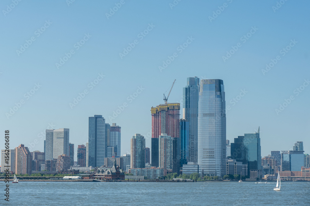 Fototapeta premium Skyline View: A cityscape panorama of modern buildings, a tower under construction, and sailboats on the water against a serene blue sky.