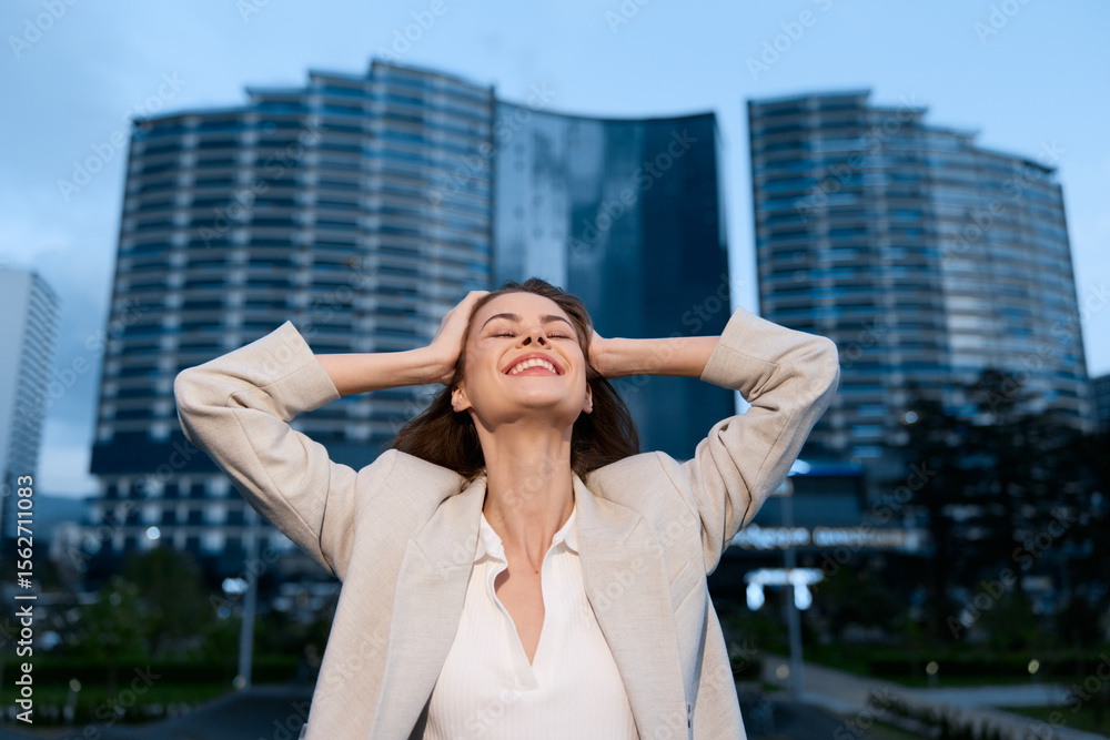 Fototapeta premium Confident businesswoman celebrating success outdoors in a modern urban setting with skyscrapers in the background, conveying professional empowerment and achievement.