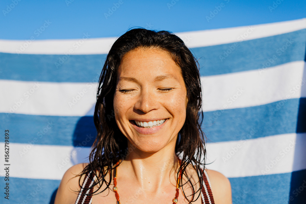 © BONNINSTUDIO/Stocksy - Happy woman smiling with eyes closed at beach with striped towel © BONNINSTUDIO/Stocksy - Happy woman smiling with eyes closed at beach with striped towel
