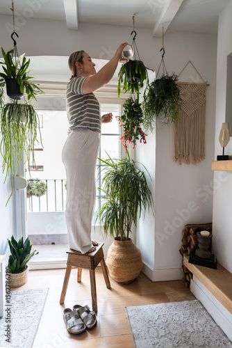 Woman watering hanging plants standing on wooden stool