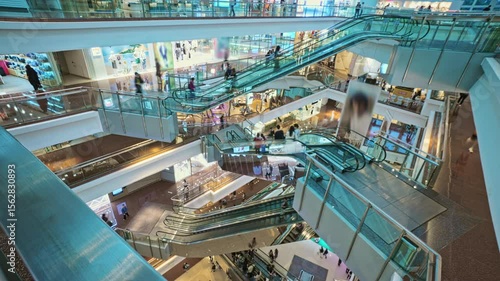 Bright Multi-Level Shopping Mall Interior Revealing Escalators Transporting Shoppers Between Floors. Very Busy Full of Clients Shopping Mall Interior With Escalators And Customers. Time Lapse
